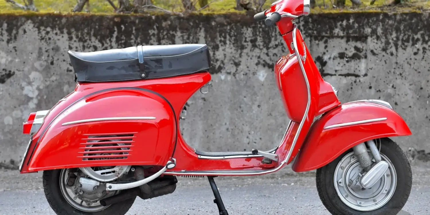 Seitenansicht einer restaurierten roten Piaggio Vespa 180 Super Sport von 1966 vor einer Steinmauer.