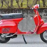Seitenansicht einer restaurierten roten Piaggio Vespa 180 Super Sport von 1966 vor einer Steinmauer.