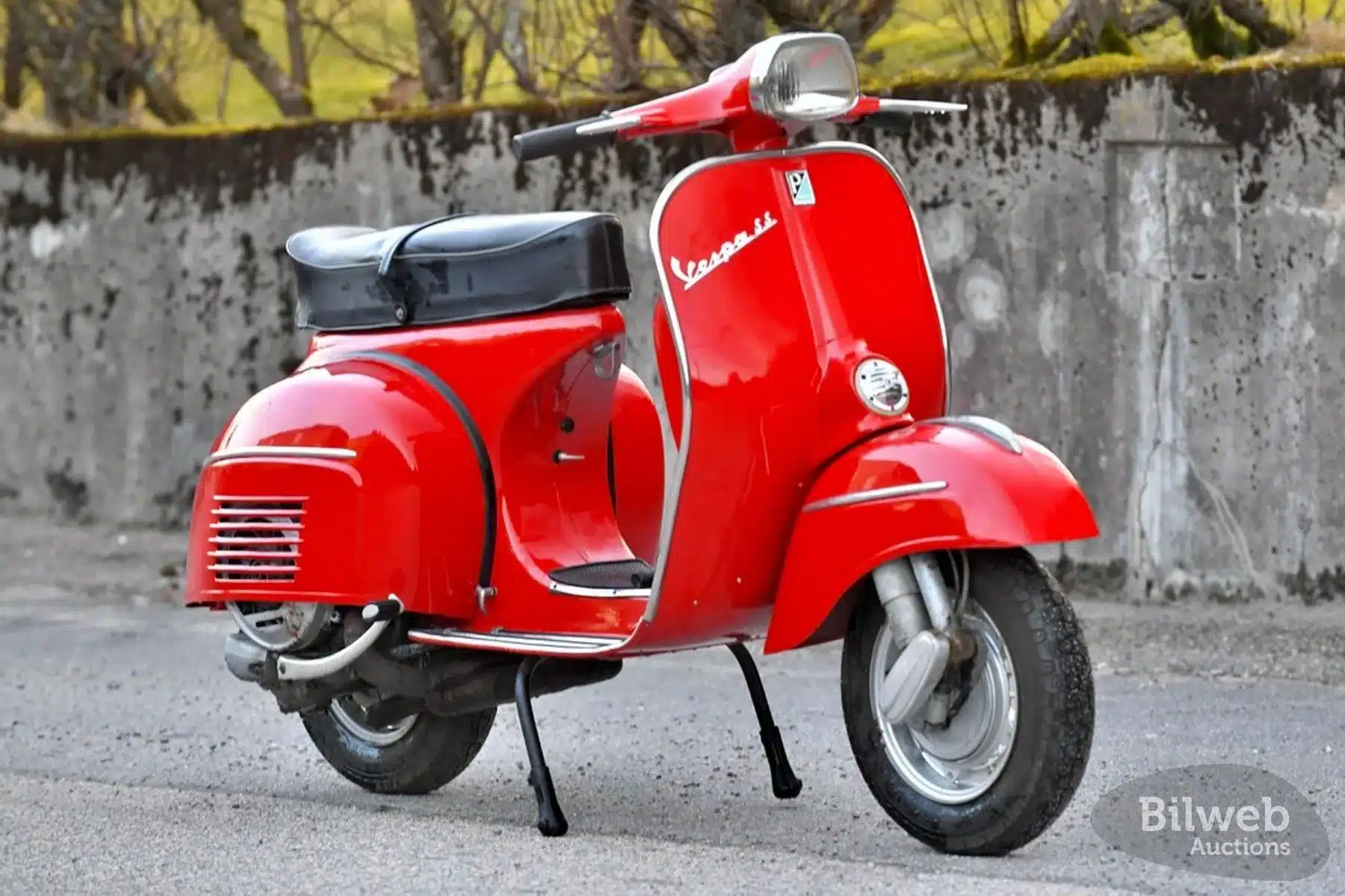 Rote Piaggio Vespa 180 Super Sport von 1966 mit schwarzer Sitzbank, seitlich und leicht von vorne vor Betonmauer stehend