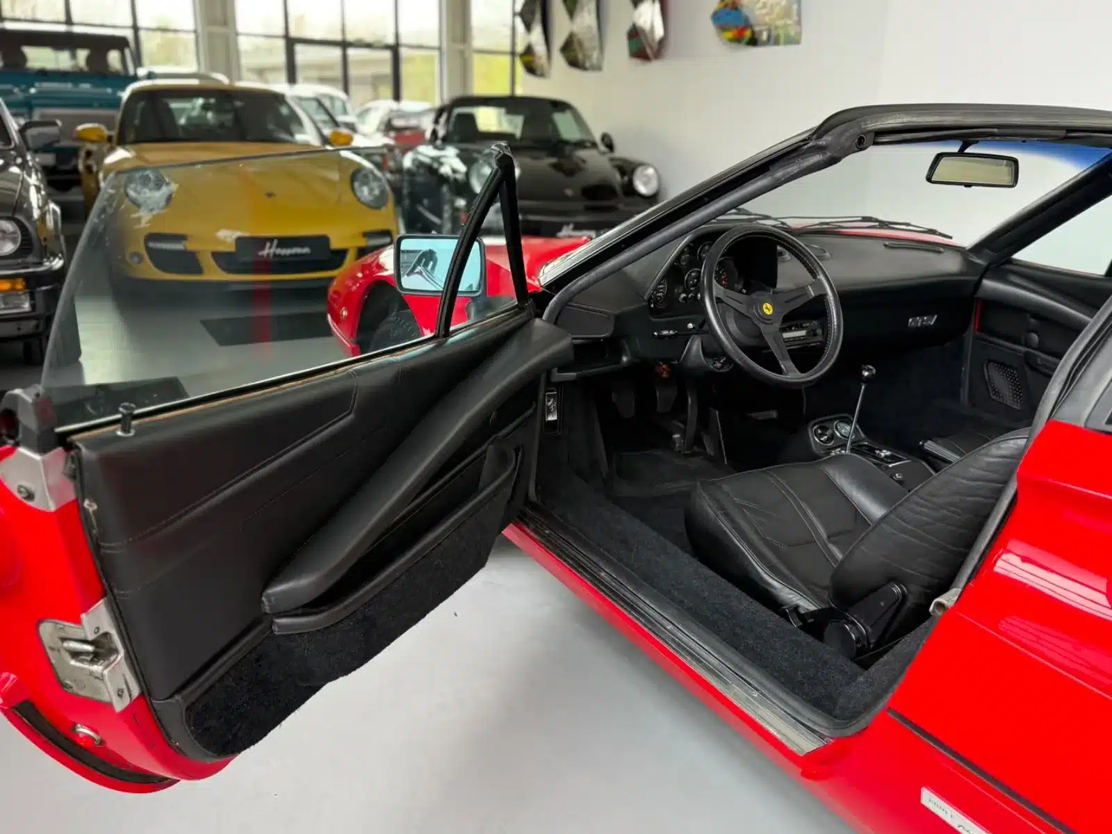 Innenraum eines Ferrari 308 GTSi mit schwarzen Ledersitzen und geöffneter Fahrertür, Blick auf das Cockpit und Lenkrad, Targa-typische Dachkonstruktion sichtbar.