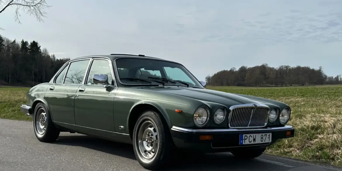 Grüner Jaguar XJ6 Series III von 1985 steht auf einer Landstraße vor ländlicher Kulisse.