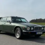 Grüner Jaguar XJ6 Series III von 1985 steht auf einer Landstraße vor ländlicher Kulisse.
