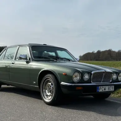 Grüner Jaguar XJ6 Series III von 1985 steht auf einer Landstraße vor ländlicher Kulisse.