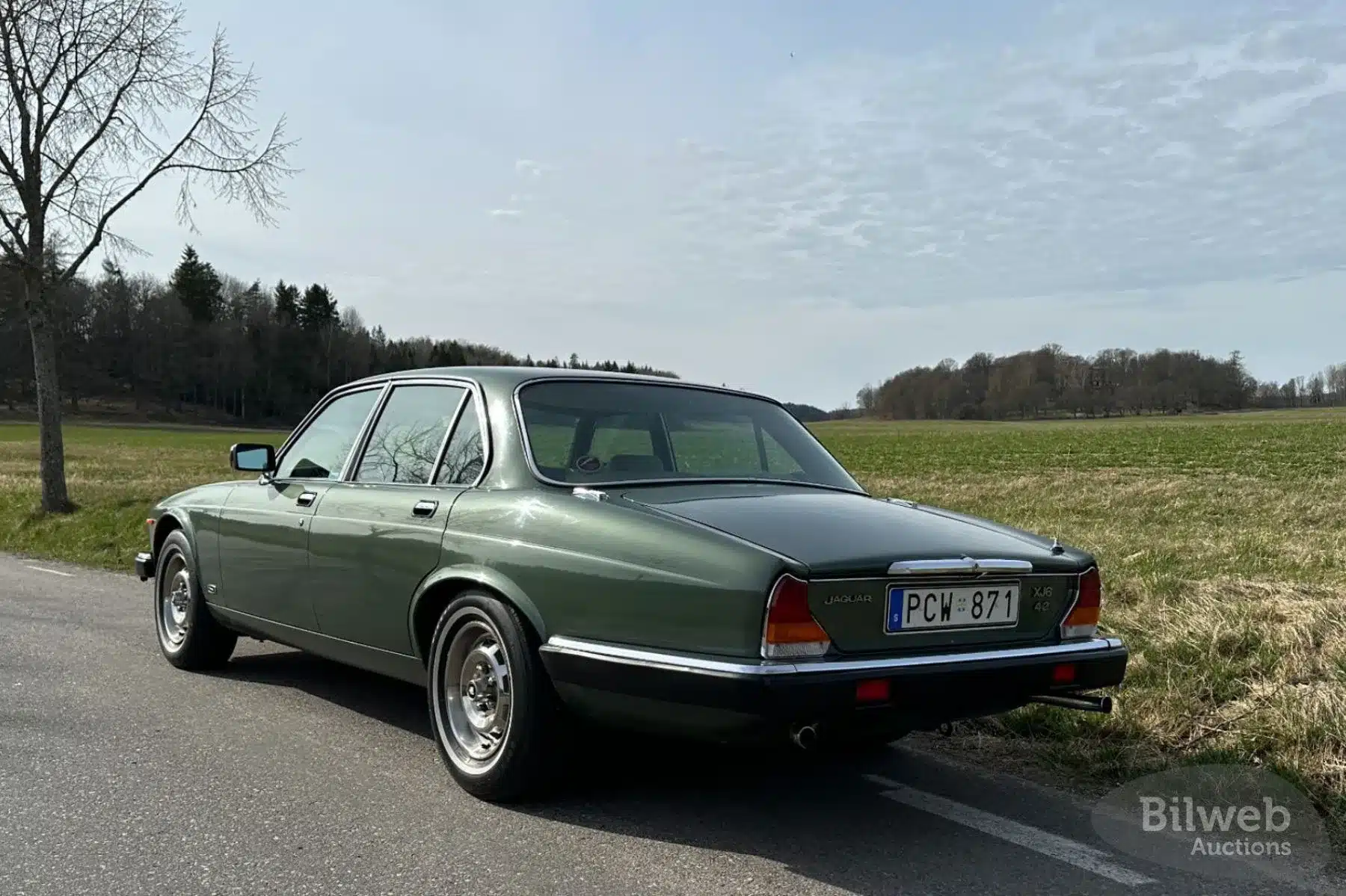 Grüner Jaguar XJ6 Series III 4.2L von 1985, Heckansicht auf einer Landstraße vor ländlicher Kulisse