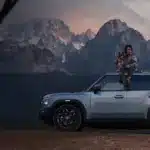 JETOUR T2 i-DM SUV mit Panoramadach, Mann mit Hund auf dem Dach, Berge im Hintergrund bei Sonnenuntergang.