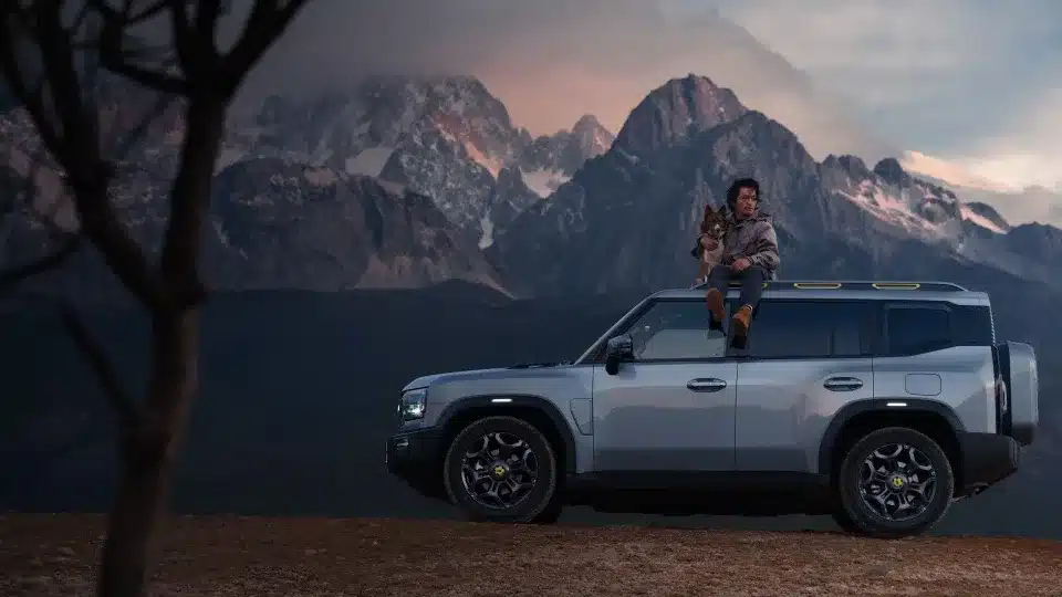 JETOUR T2 i-DM SUV mit Panoramadach, Mann mit Hund auf dem Dach, Berge im Hintergrund bei Sonnenuntergang.