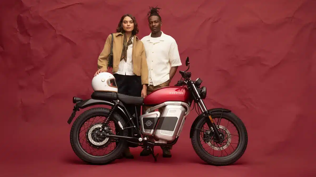 Elektromotorrad Maeving RM2 mit zwei stehenden Personen und Motorradhelm vor rotem Hintergrund