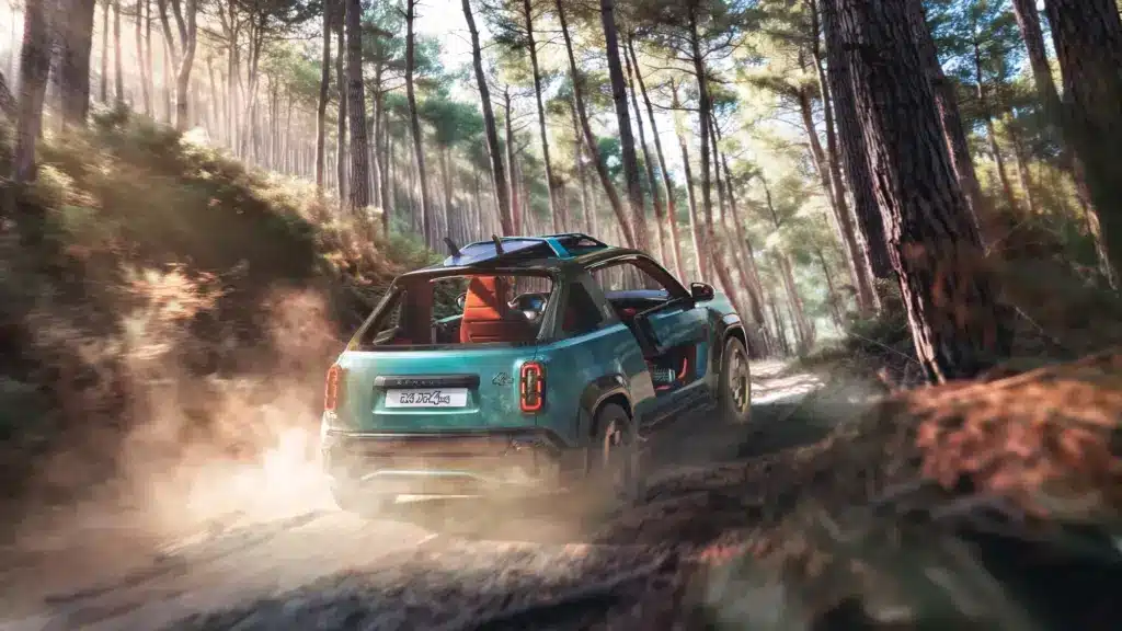 Renault 4 JP4x4 Concept in türkisgrün fährt mit geöffnetem Dach und Surfbrett auf dem Dachträger durch einen lichten Wald auf einer staubigen Piste.