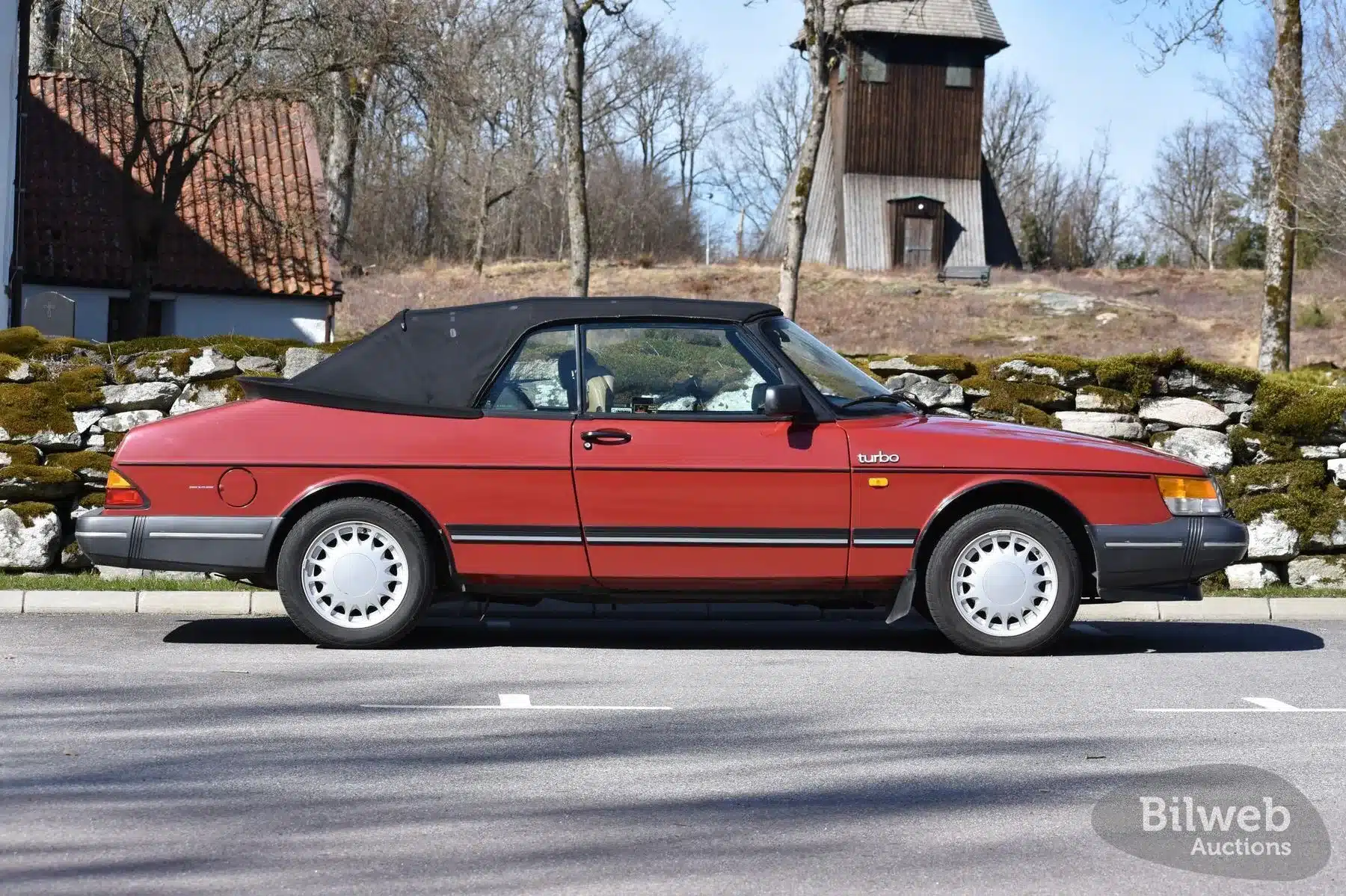 Rotes Saab 900 T16 Cabriolet 1988 mit geschlossenem Verdeck, Seitenansicht, aufgenommen auf einem Parkplatz vor einer Steinmauer und Bäumen.