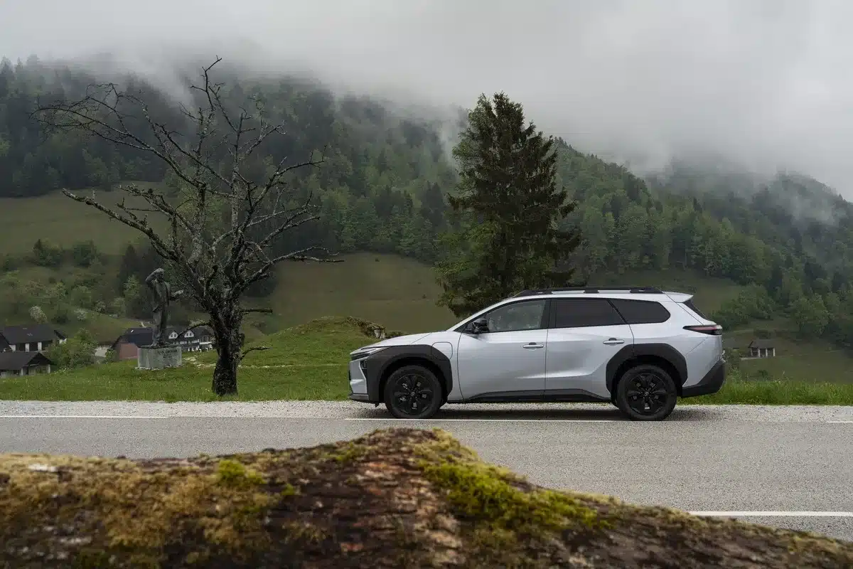 Seitenansicht eines silbernen Toyota bZ4X Touring auf einer Alpinstraße mit Bäumen und nebligen Bergen im Hintergrund.