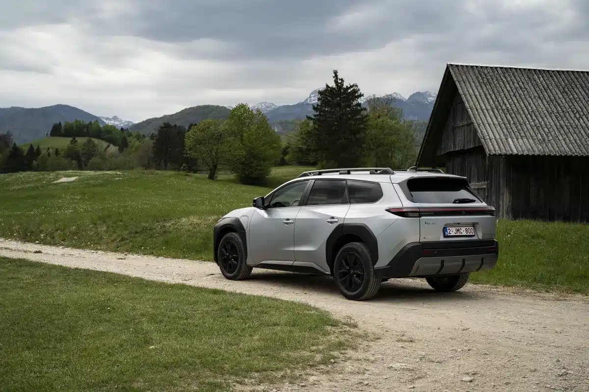 Silberner Toyota bZ4X Touring steht auf einem Feldweg vor einer Holzhütte mit Bergen im Hintergrund.