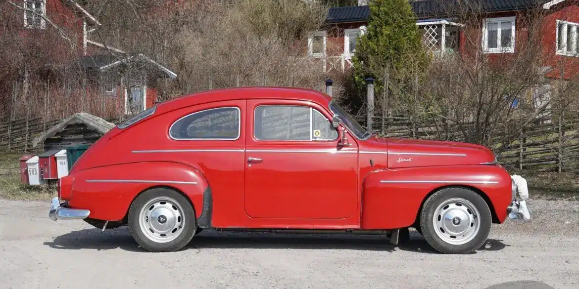 Roter Volvo PV 544 Sport B18B von 1962, seitlich aufgenommen, geparkt auf einer Schotterstraße vor ländlichen Häusern.
