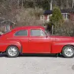 Roter Volvo PV 544 Sport B18B von 1962, seitlich aufgenommen, geparkt auf einer Schotterstraße vor ländlichen Häusern.