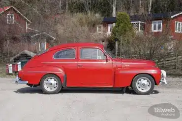 Roter Volvo PV 544 Sport B18B von 1962, seitlich aufgenommen, geparkt auf einer Schotterstraße vor ländlichen Häusern.
