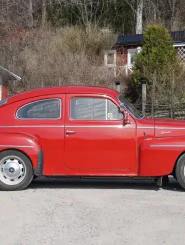 Roter Volvo PV 544 Sport B18B von 1962, seitlich aufgenommen, geparkt auf einer Schotterstraße vor ländlichen Häusern.