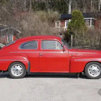 Roter Volvo PV 544 Sport B18B von 1962, seitlich aufgenommen, geparkt auf einer Schotterstraße vor ländlichen Häusern.