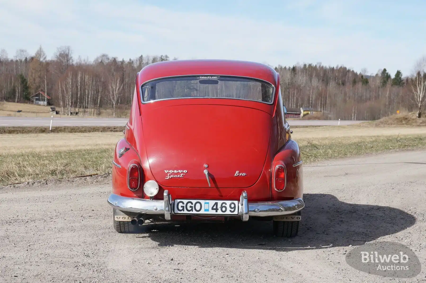 Rote Heckansicht eines Volvo PV 544 Sport B18B von 1962 auf einer Schotterstraße, Nummernschild sichtbar, ländliche Umgebung im Hintergrund.