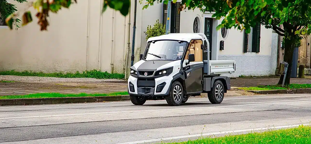 Elektrischer Alke ATX 440S fährt auf städtischer Straße vor Gebäuden, kompaktes Arbeitsfahrzeug mit Ladefläche.