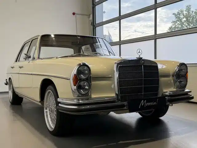 Mercedes-Benz 250 SE W108 in Beige, tiefergelegt mit BBS-Leichtmetallrädern, seitlich von vorne fotografiert in einer modernen Halle.
