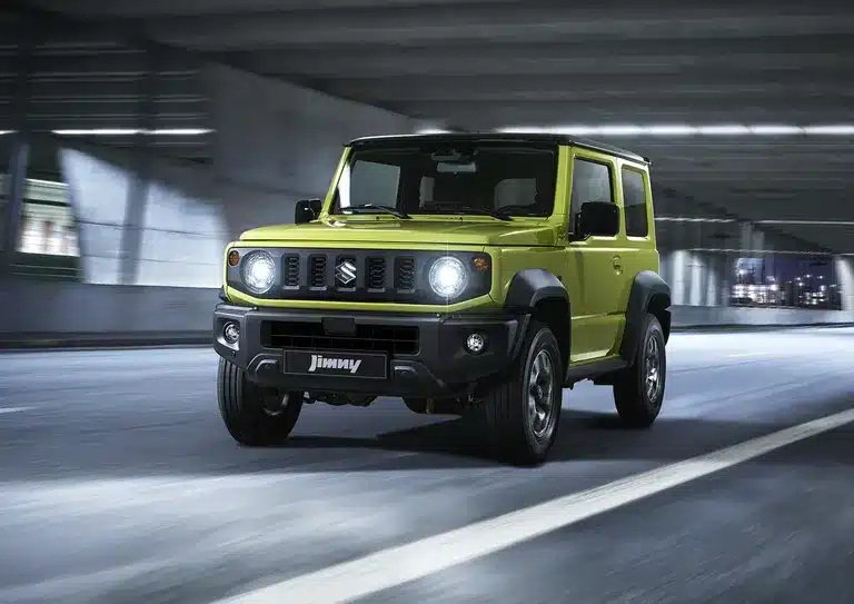 Suzuki Jimny Dreitürer in einer urbanen Umgebung, vorne links, fahrend, in hellgrüner Lackierung.