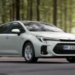Weißer Suzuki Swace Hybrid fährt auf einer Straße durch einen Wald, Seitenansicht, Tageslicht.