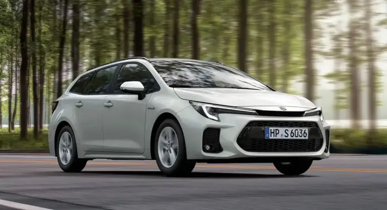 Weißer Suzuki Swace Hybrid fährt auf einer Straße durch einen Wald, Seitenansicht, Tageslicht.