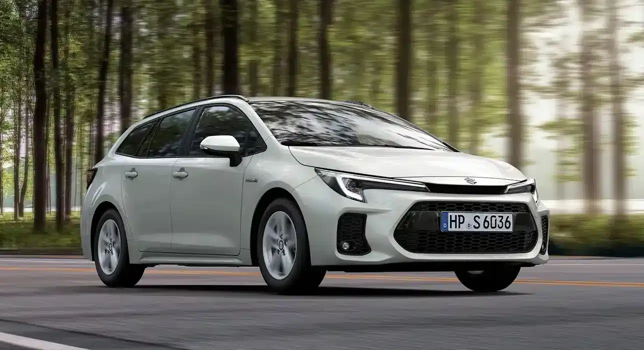 Weißer Suzuki Swace Hybrid fährt auf einer Straße durch einen Wald, Seitenansicht, Tageslicht.