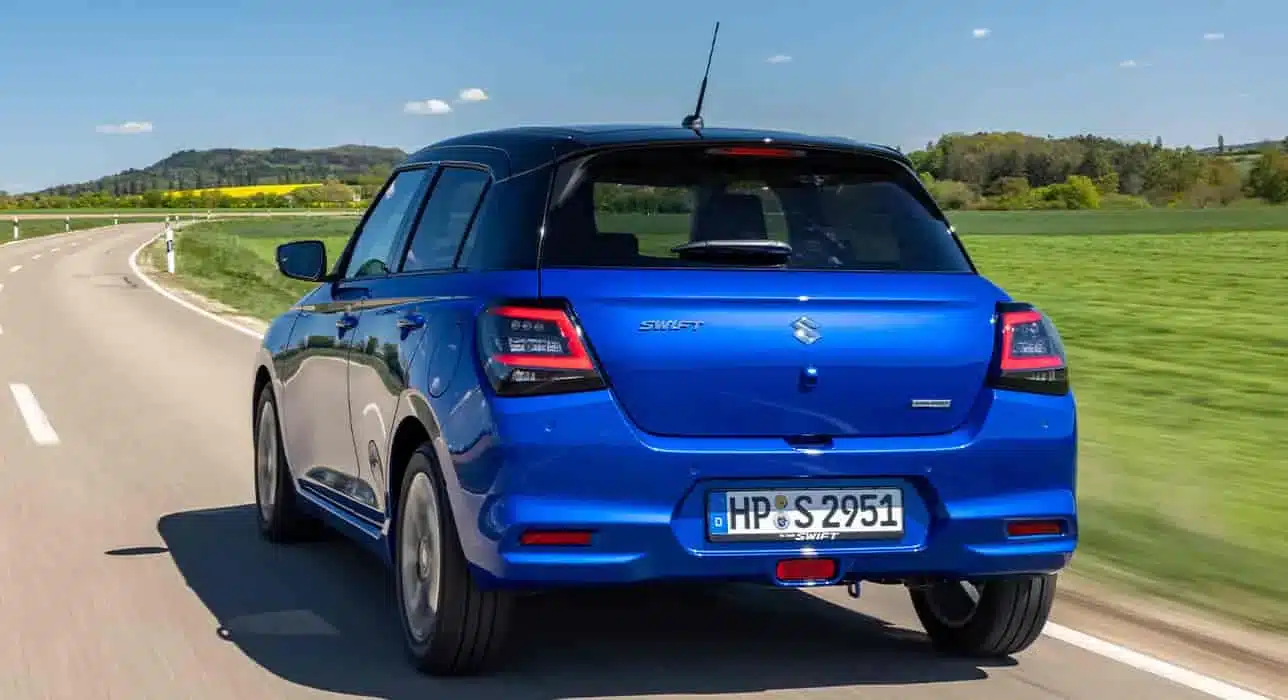 Blauer Suzuki Swift (2024) fährt auf einer Landstraße, Rückansicht mit Heckleuchten und serienmäßigem Dachspoiler.