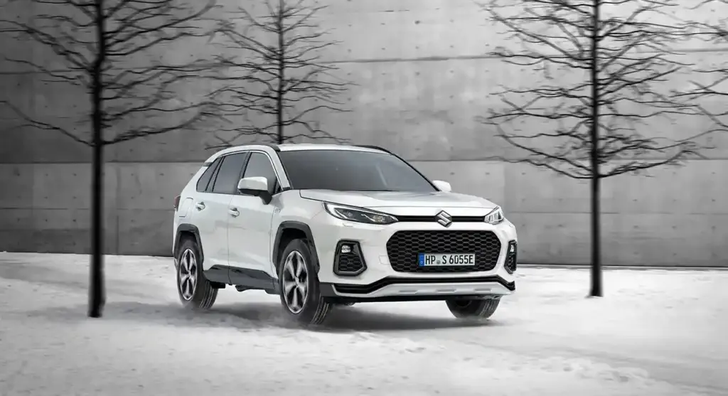 Weißer Suzuki Across Plug-in-Hybrid SUV auf verschneitem Untergrund vor einer Betonwand mit kahlen Bäumen.