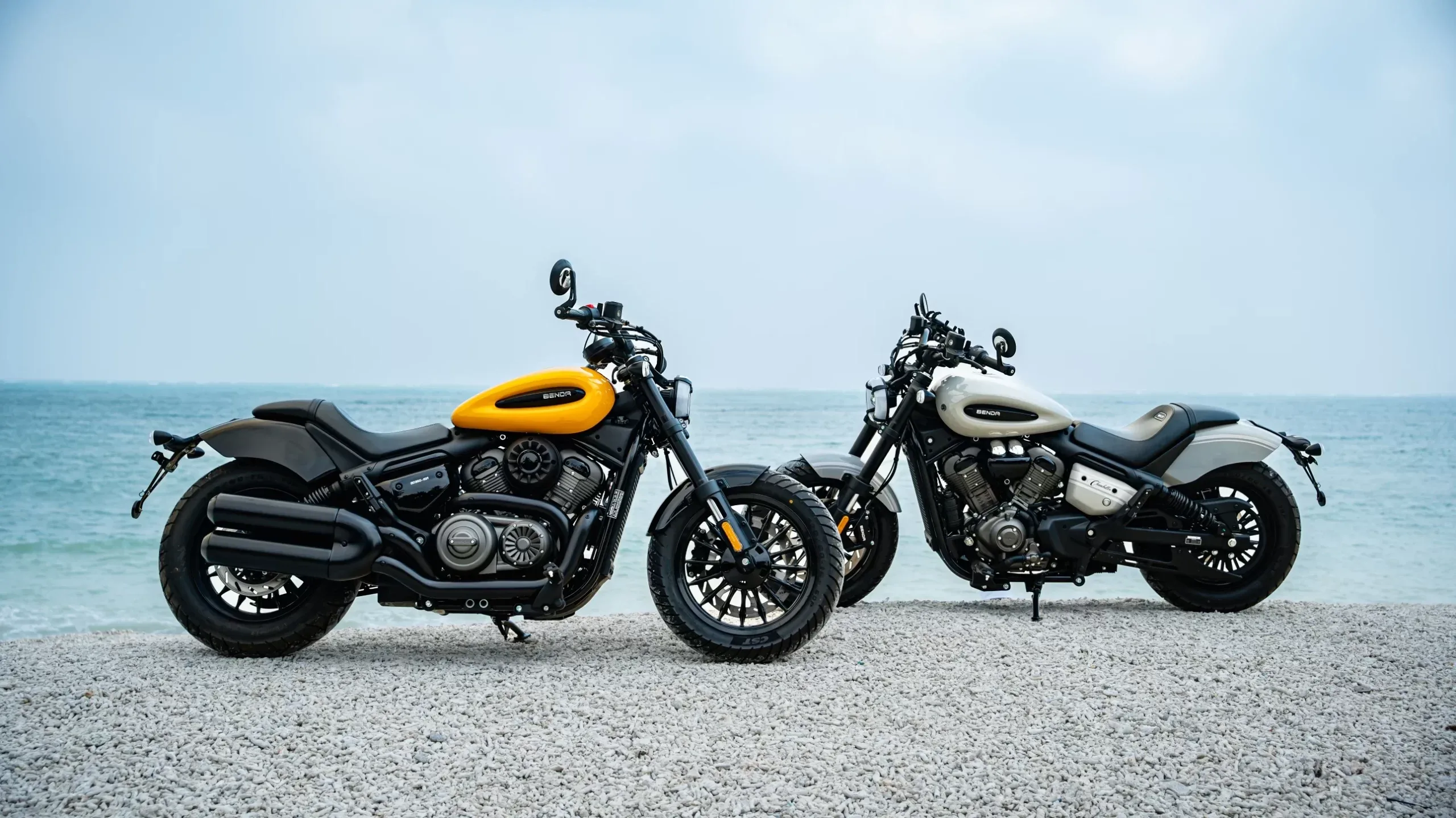 Zwei Chinchilla 350 CVT NEO Motorräder, eines gelb, eines weiß, stehen nebeneinander am Kiesstrand mit Blick aufs Wasser.