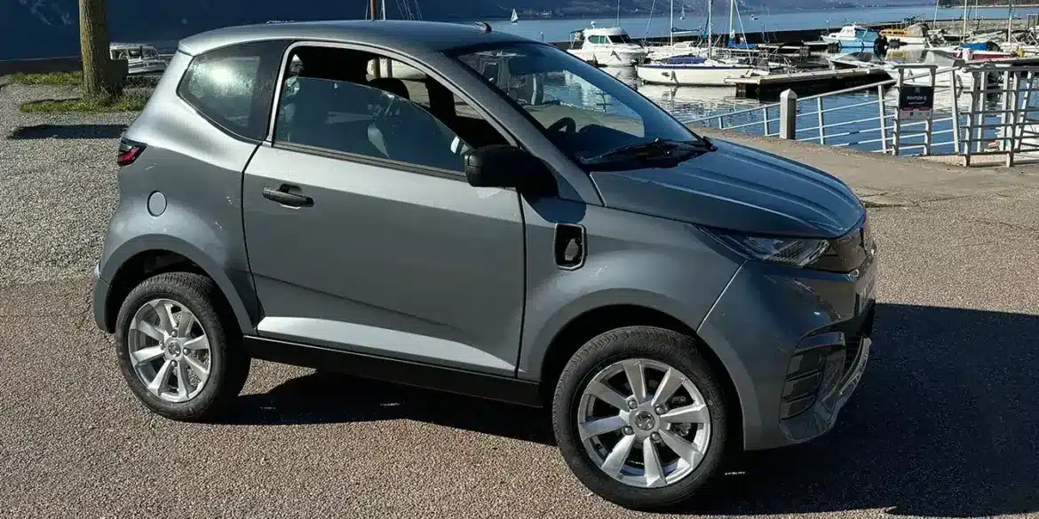 Aixam E City Pack in silbermetallic, dreitüriges leichtes Elektroauto auf einem Parkplatz am Wasser, Seitenansicht.