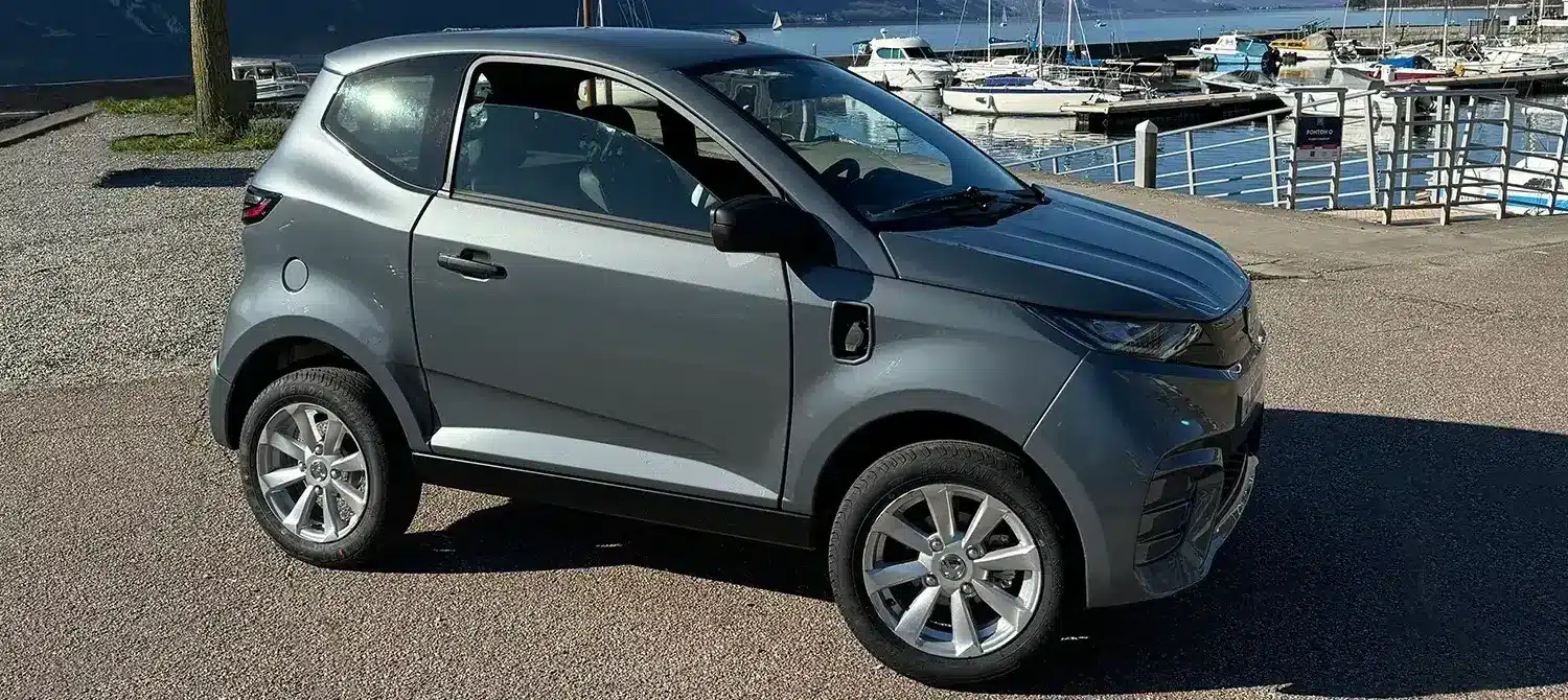 Aixam E City Pack in silbermetallic, dreitüriges leichtes Elektroauto auf einem Parkplatz am Wasser, Seitenansicht.