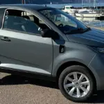 Aixam E City Pack in silbermetallic, dreitüriges leichtes Elektroauto auf einem Parkplatz am Wasser, Seitenansicht.