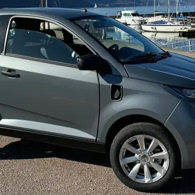 Aixam E City Pack in silbermetallic, dreitüriges leichtes Elektroauto auf einem Parkplatz am Wasser, Seitenansicht.