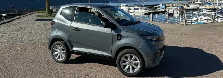 Aixam E City Pack in silbermetallic, dreitüriges leichtes Elektroauto auf einem Parkplatz am Wasser, Seitenansicht.