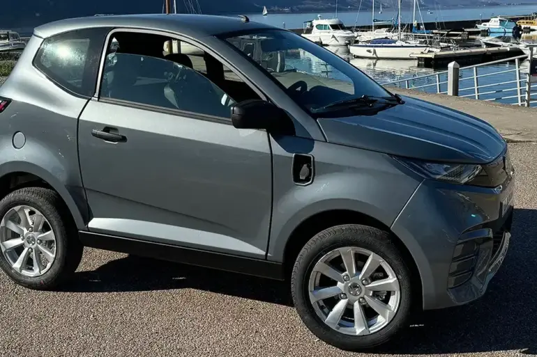 Aixam E City Pack in silbermetallic, dreitüriges leichtes Elektroauto auf einem Parkplatz am Wasser, Seitenansicht.