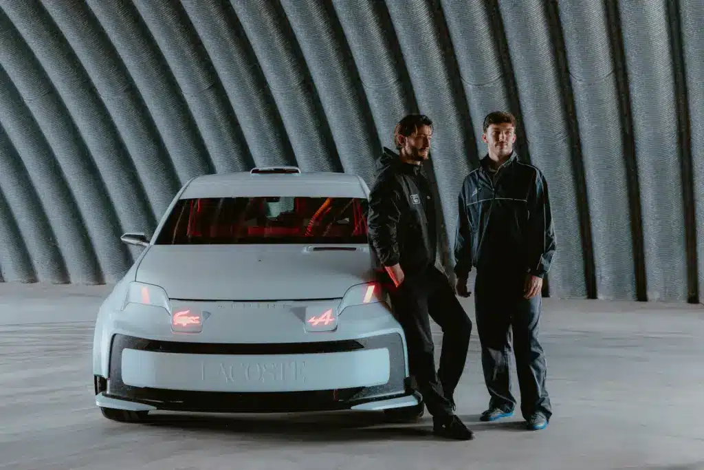 Alpine Lacoste A290 Rallye und zwei Personen in einem modernen Hangar, Frontansicht des Fahrzeugs mit Logos.
