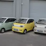 Drei ARI Bruni Elektroautos nebeneinander, pastellgrün, gelb und weiß, vor Garagentoren