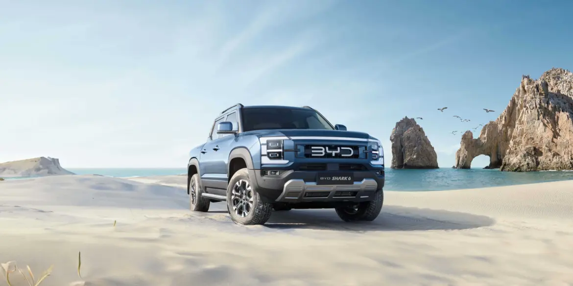BYD Shark 6 Pickup fährt über einen sonnigen Sandstrand mit Felsen im Hintergrund.