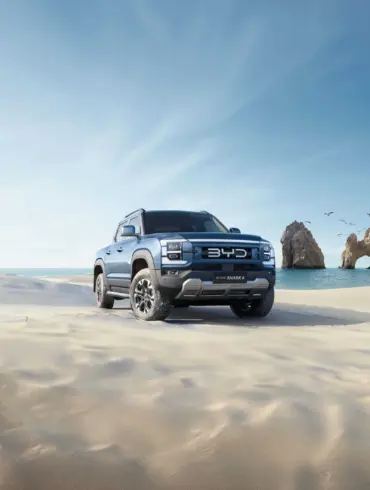 BYD Shark 6 Pickup fährt über einen sonnigen Sandstrand mit Felsen im Hintergrund.