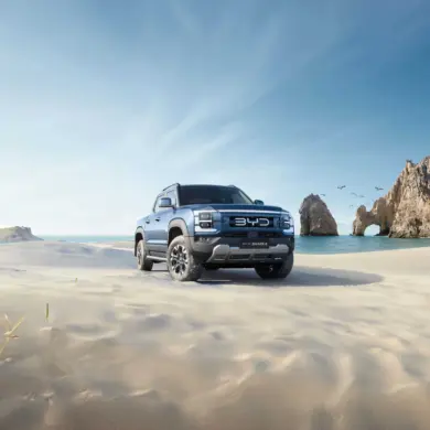 BYD Shark 6 Pickup fährt über einen sonnigen Sandstrand mit Felsen im Hintergrund.