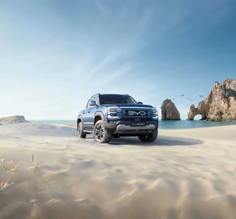 BYD Shark 6 Pickup fährt über einen sonnigen Sandstrand mit Felsen im Hintergrund.