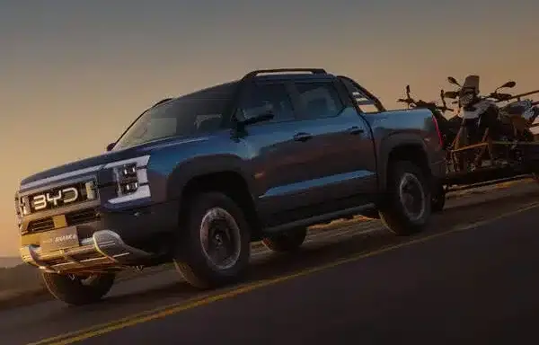 BYD Shark 6 fährt mit Motorrad-Anhänger auf einer Landstraße bei Sonnenuntergang
