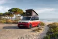 VW California Ocean „Generation“ in Candy-Weiß/Sunset Red Metallic mit Aufstelldach am Strand geparkt