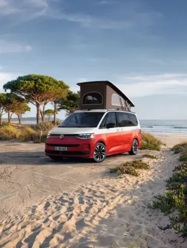 VW California Ocean „Generation“ in Candy-Weiß/Sunset Red Metallic mit Aufstelldach am Strand geparkt