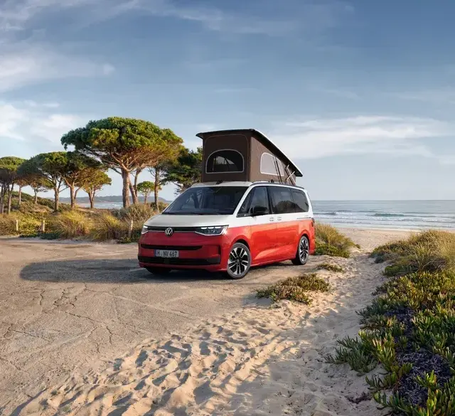 VW California Ocean „Generation“ in Candy-Weiß/Sunset Red Metallic mit Aufstelldach am Strand geparkt