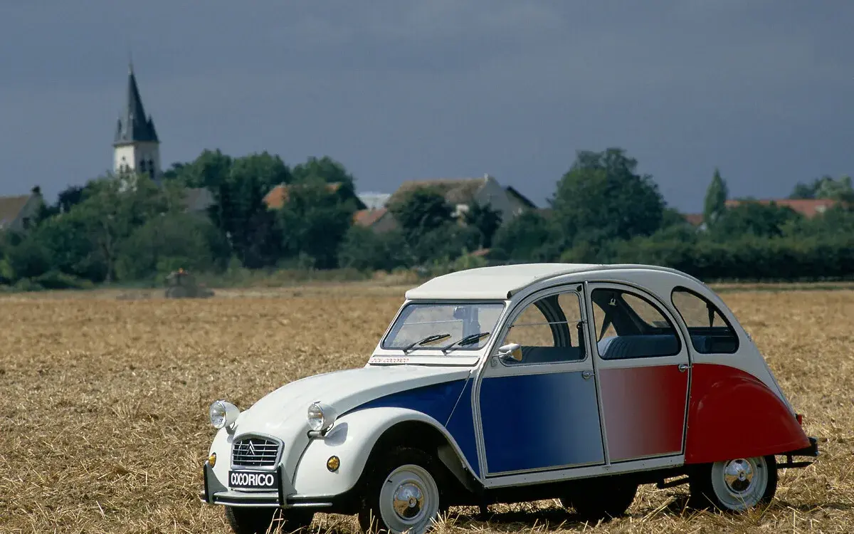 Citroën 2CV Cocorico Sonderedition in Blau, Weiß und Rot auf einem Stoppelfeld, seitliche Ansicht, ländliche Kulisse.