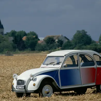 Citroën 2CV Cocorico Sonderedition in Blau, Weiß und Rot auf einem Stoppelfeld, seitliche Ansicht, ländliche Kulisse.