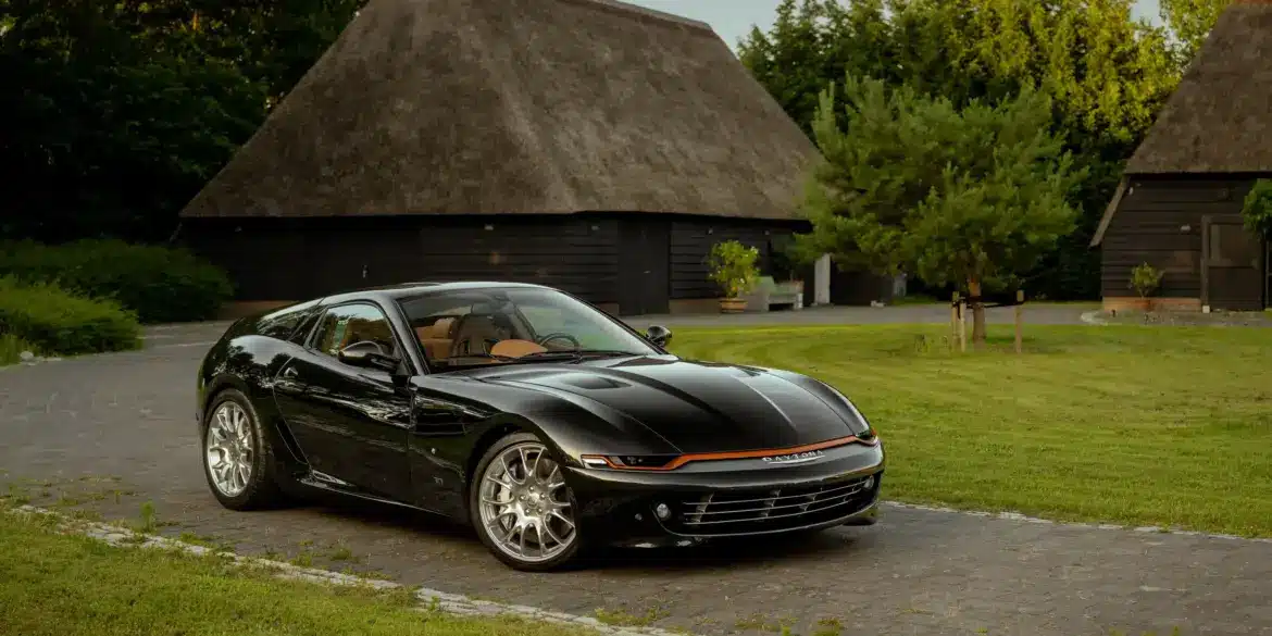 Schwarzer Daytona Shooting Brake Hommage parkt auf einer Auffahrt vor traditionellen strohgedeckten Gebäuden.