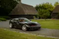 Schwarzer Daytona Shooting Brake Hommage parkt auf einer Auffahrt vor traditionellen strohgedeckten Gebäuden.